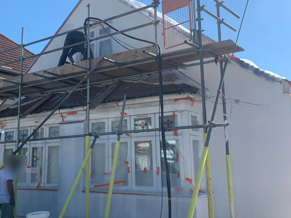 Chalet bungalow in Dagenham under scaffolding, with the bay windows sheeted and the side wall freshly rendered white