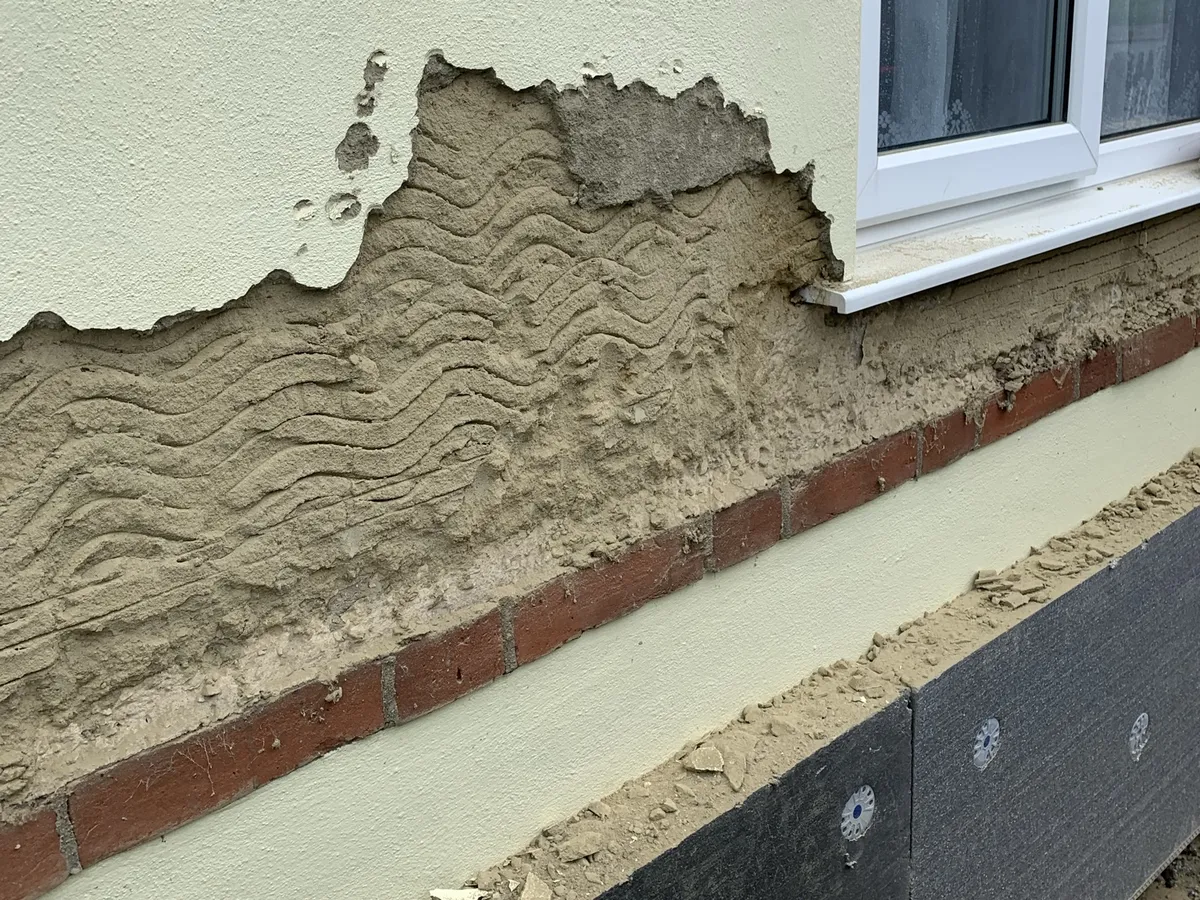 Old render coming away from a bungalow wall below a window, with insulation boards fixed along the base