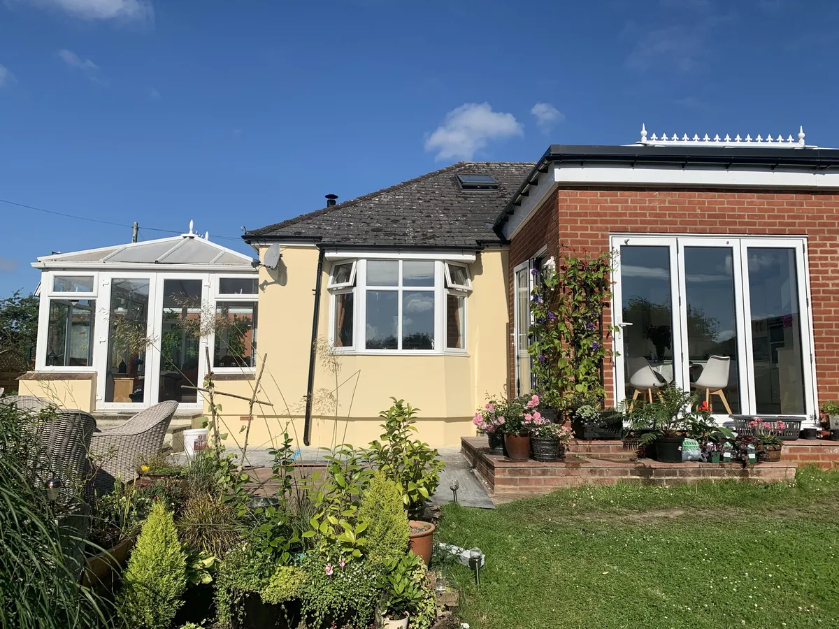 Rear of the same Hertfordshire bungalow finished in pale yellow render, with a conservatory and a brick extension