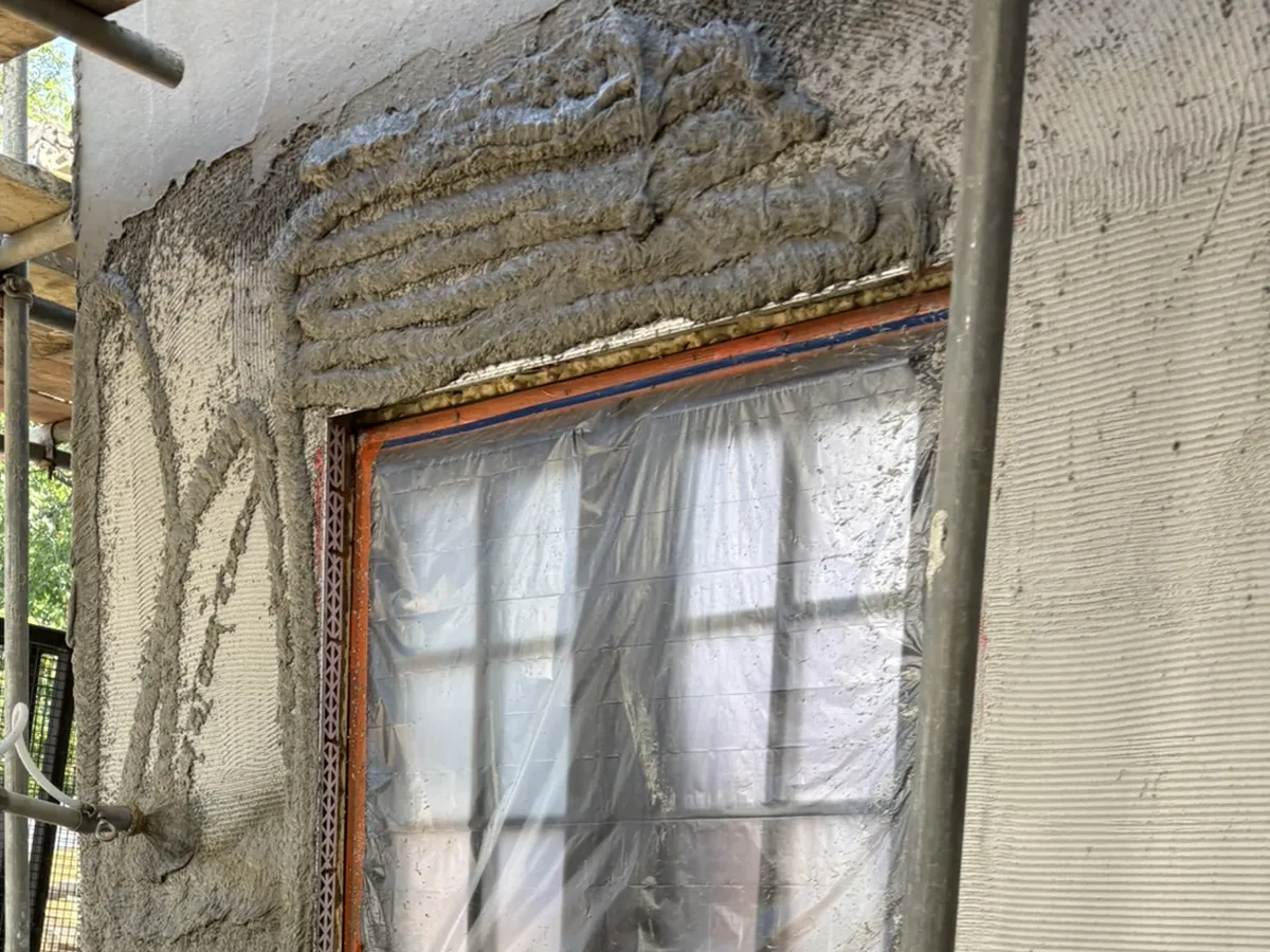 Render being sprayed onto a wall by machine around a masked window, next to a section already ruled flat